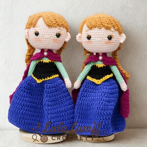 Puede incluir: Dos mu&ntilde;ecas de ganchillo hechas a mano, cada una con cabello casta&ntilde;o rojizo en trenzas, vestidas con un vestido azul, un corpi&ntilde;o negro y una capa morada. Las mu&ntilde;ecas se exhiben sobre bases de madera. El texto "Lala Lamb Crochet" es visible.