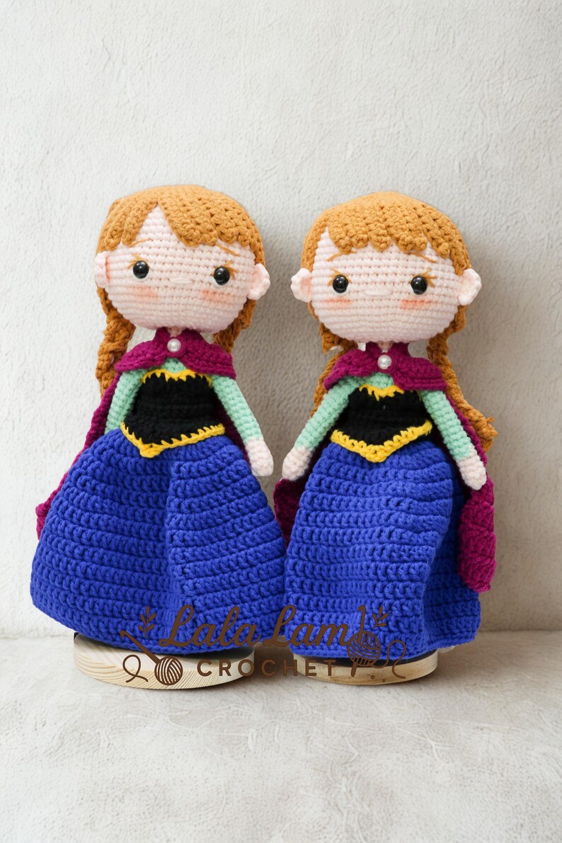 Puede incluir: Dos mu&ntilde;ecas de ganchillo hechas a mano, cada una con cabello casta&ntilde;o rojizo en trenzas, vestidas con un vestido azul, un corpi&ntilde;o negro y una capa morada. Las mu&ntilde;ecas se exhiben sobre bases de madera. El texto "Lala Lamb Crochet" es visible.