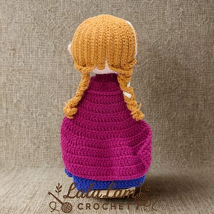 Puede incluir: Mu&ntilde;eca de ganchillo hecha a mano con peinado trenzado amarillo dorado, que lleva una capa magenta y un vestido azul. La mu&ntilde;eca est&aacute; hecha con puntadas visibles, mostrando una apariencia texturizada.