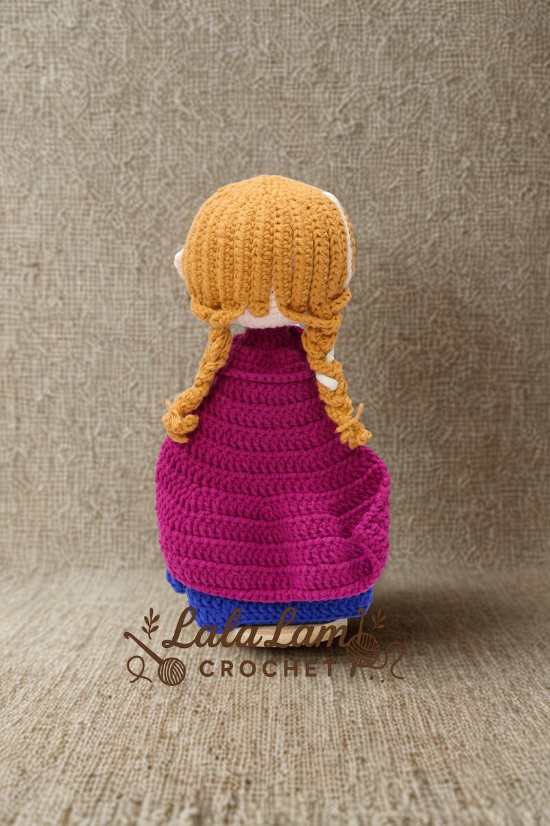 Puede incluir: Mu&ntilde;eca de ganchillo hecha a mano con peinado trenzado amarillo dorado, que lleva una capa magenta y un vestido azul. La mu&ntilde;eca est&aacute; hecha con puntadas visibles, mostrando una apariencia texturizada.