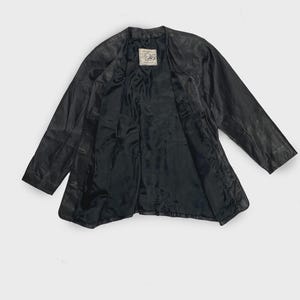 May include: Black leather jacket with a classic design. The jacket has a V-neck collar and a button closure. The interior lining is black. A label is visible on the inside of the jacket. The jacket is spread out on a white background.