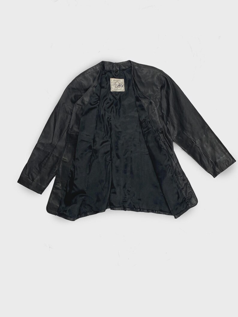 May include: Black leather jacket with a classic design. The jacket has a V-neck collar and a button closure. The interior lining is black. A label is visible on the inside of the jacket. The jacket is spread out on a white background.