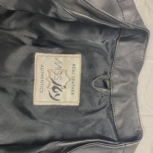 May include: Black leather jacket with a label that reads "Miss Joy Authentics Real Leather". The jacket is partially unzipped, revealing the interior lining. The leather appears to be slightly worn.