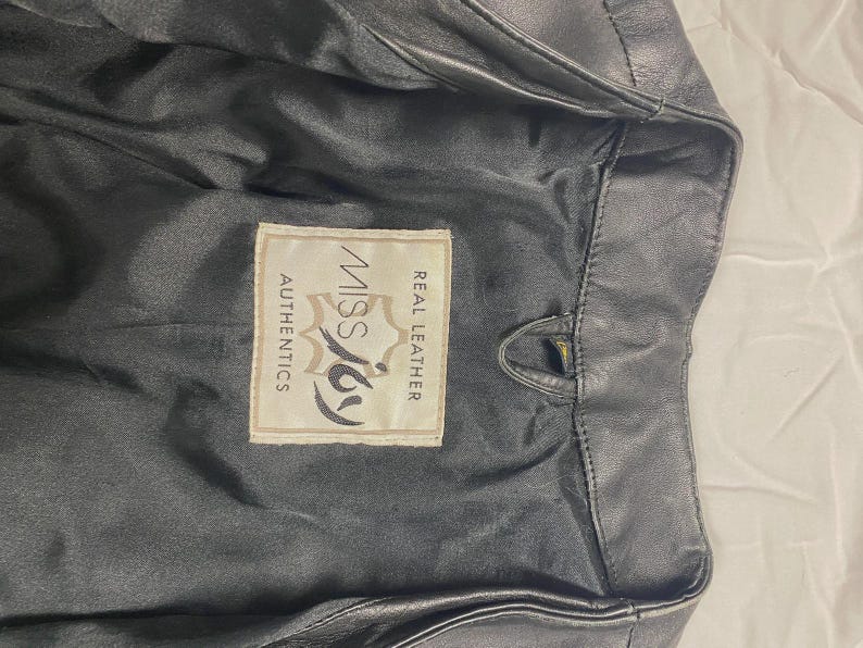 May include: Black leather jacket with a label that reads "Miss Joy Authentics Real Leather". The jacket is partially unzipped, revealing the interior lining. The leather appears to be slightly worn.