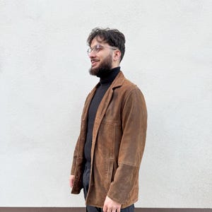 K&ouml;nnte beinhalten: Ein brauner Wildlederblazer mit klassischem Design. Die Jacke hat ein fallendes Revers, einen Knopfverschluss und aufgesetzte Taschen. Der Blazer wird &uuml;ber einem schwarzen Rollkragenpullover und mit einer schwarzen Hose getragen.