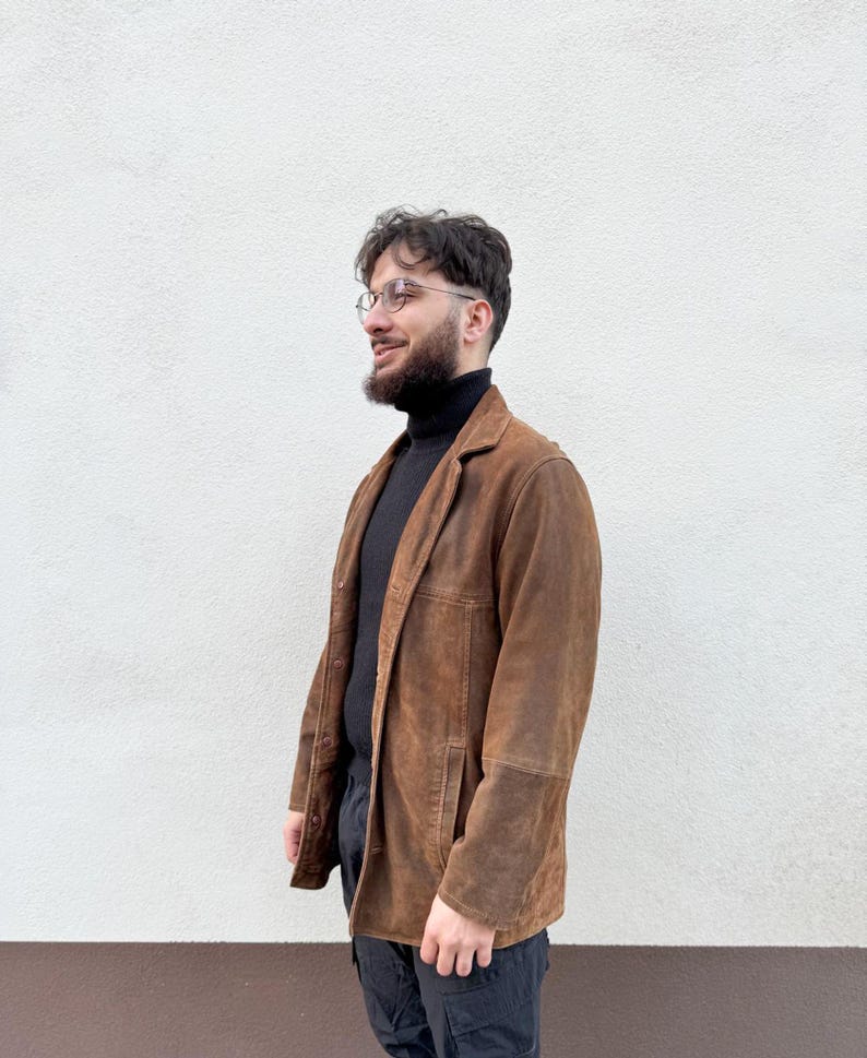 K&ouml;nnte beinhalten: Ein brauner Wildlederblazer mit klassischem Design. Die Jacke hat ein fallendes Revers, einen Knopfverschluss und aufgesetzte Taschen. Der Blazer wird &uuml;ber einem schwarzen Rollkragenpullover und mit einer schwarzen Hose getragen.