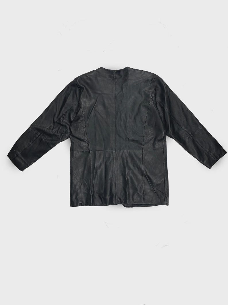 May include: A black leather jacket with a round neckline and long sleeves. The jacket is laid flat, showcasing its tailored design and seams. The leather appears slightly textured.
