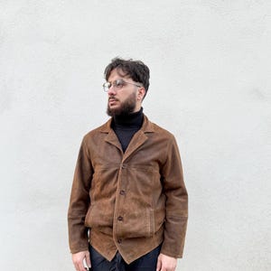 K&ouml;nnte beinhalten: Braune Wildlederjacke mit Knopfleiste und zwei Vordertaschen. Die Jacke hat einen klassischen Kragen und eine taillierte Passform. Die Person tr&auml;gt einen schwarzen Rollkragenpullover und eine schwarze Hose. Der Hintergrund ist eine schlichte wei&szlig;e Wand.
