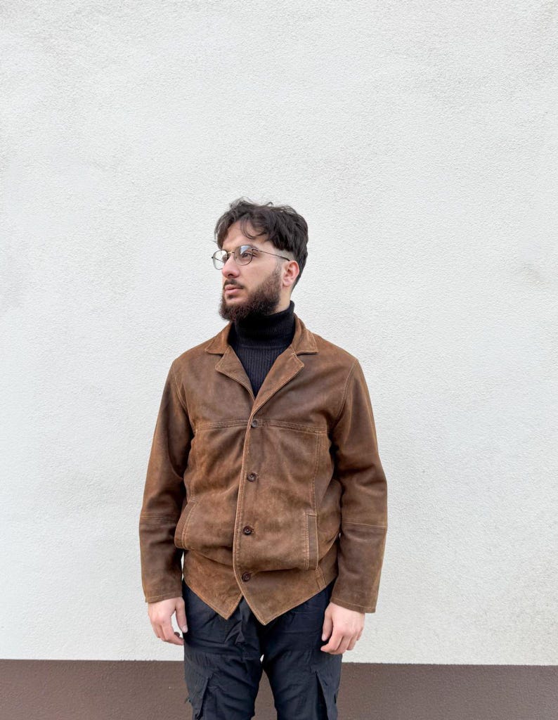 K&ouml;nnte beinhalten: Braune Wildlederjacke mit Knopfleiste und zwei Vordertaschen. Die Jacke hat einen klassischen Kragen und eine taillierte Passform. Die Person tr&auml;gt einen schwarzen Rollkragenpullover und eine schwarze Hose. Der Hintergrund ist eine schlichte wei&szlig;e Wand.