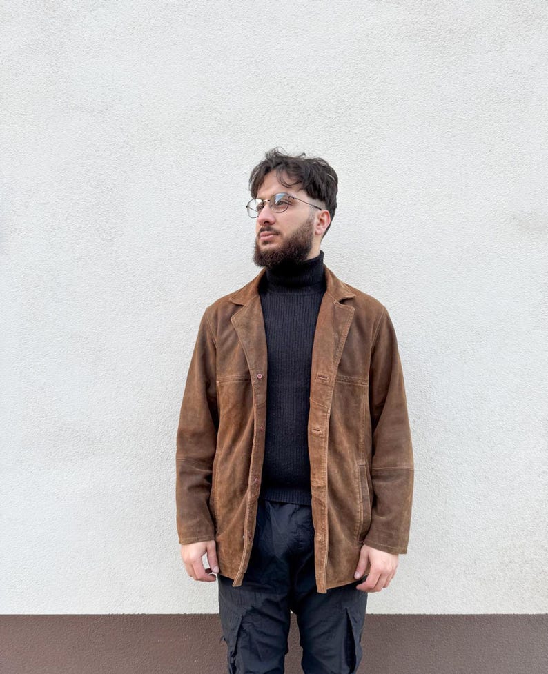 K&ouml;nnte beinhalten: Braune Wildleder-Blazer mit klassischem Kragen und Knopfverschluss. Die Jacke wird &uuml;ber einem schwarzen Rollkragenpullover und mit schwarzen Cargohosen getragen. Der Hintergrund ist eine schlichte wei&szlig;e Wand.