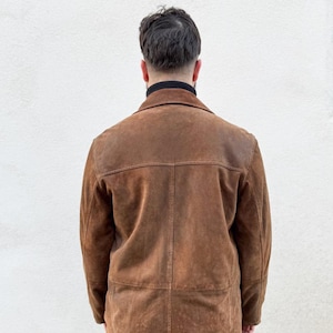 K&ouml;nnte beinhalten: Braune Wildlederjacke mit klassischem Kragen und taillierter Passform. Die Jacke hat ein abgenutztes, strukturiertes Aussehen und einen Knopfverschluss. Das Model tr&auml;gt einen schwarzen Rollkragenpullover darunter. Die Jacke ist im Vintage-Stil.
