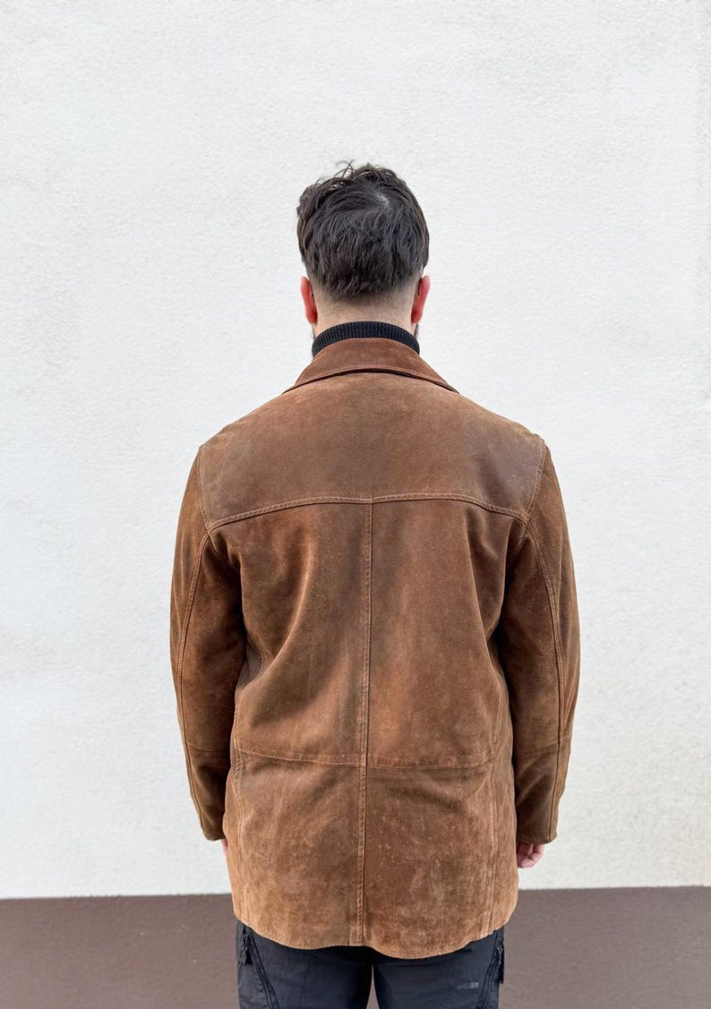 K&ouml;nnte beinhalten: Braune Wildlederjacke mit klassischem Kragen und taillierter Passform. Die Jacke hat ein abgenutztes, strukturiertes Aussehen und einen Knopfverschluss. Das Model tr&auml;gt einen schwarzen Rollkragenpullover darunter. Die Jacke ist im Vintage-Stil.