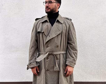 May include: A long, double-breasted trench coat in a muted olive-green tone, featuring a wide collar, shoulder epaulets, a matching waist belt with a buckle, and large buttons. The coat is worn over a black turtleneck sweater and dark trousers.
