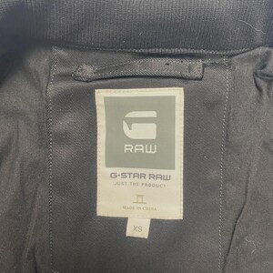 May include: Close-up of the inside of a black jacket. The label reads "G-STAR RAW JUST THE PRODUCT" with the brand's logo and "MADE IN CHINA" text. The size is XS.