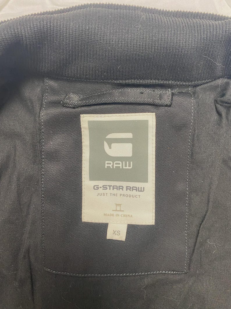 May include: Close-up of the inside of a black jacket. The label reads "G-STAR RAW JUST THE PRODUCT" with the brand's logo and "MADE IN CHINA" text. The size is XS.