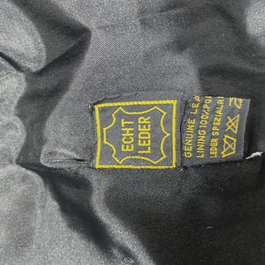 May include: Close-up of a black garment interior with a yellow and black tag. The tag reads "ECHT LEDER" in a gold frame, with additional text indicating genuine leather and lining composition.