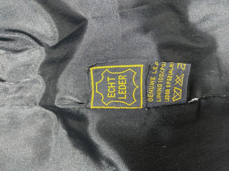 May include: Close-up of a black garment interior with a yellow and black tag. The tag reads "ECHT LEDER" in a gold frame, with additional text indicating genuine leather and lining composition.