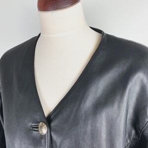 May include: A black leather jacket with a V-neckline and a single, decorative button. The jacket is displayed on a mannequin, showcasing its sleek design and tailored fit. The leather appears smooth and the jacket has a classic, timeless style.