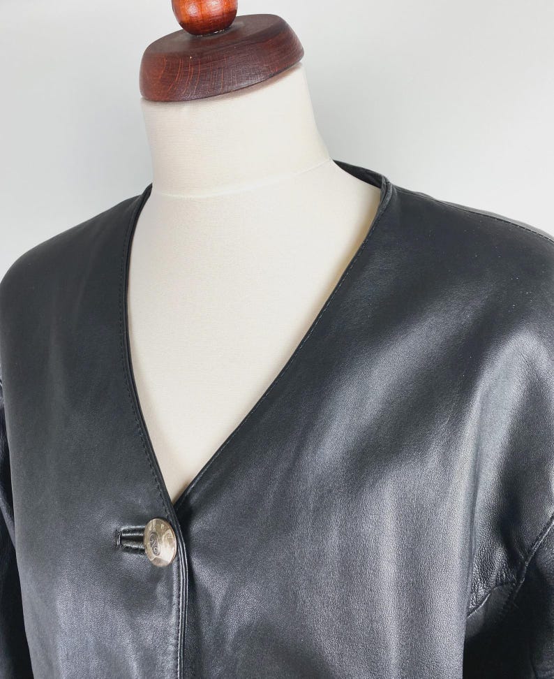 May include: A black leather jacket with a V-neckline and a single, decorative button. The jacket is displayed on a mannequin, showcasing its sleek design and tailored fit. The leather appears smooth and the jacket has a classic, timeless style.