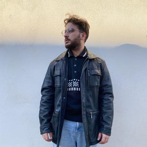 Könnte beinhalten: Schwarze Lederjacke mit mehreren Taschen und einem Kragen. Die Jacke wird über einem schwarzen Poloshirt mit einem schwarz-weißen Karomuster getragen. Die Person trägt eine blaue Jeans und eine Brille.