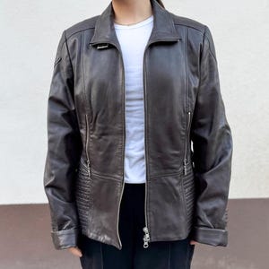 May include: A dark brown leather jacket with a zip-up front, a collar, and side pockets. The jacket features a quilted design on the lower sides and is worn over a white t-shirt. The sleeves are cuffed at the wrists.