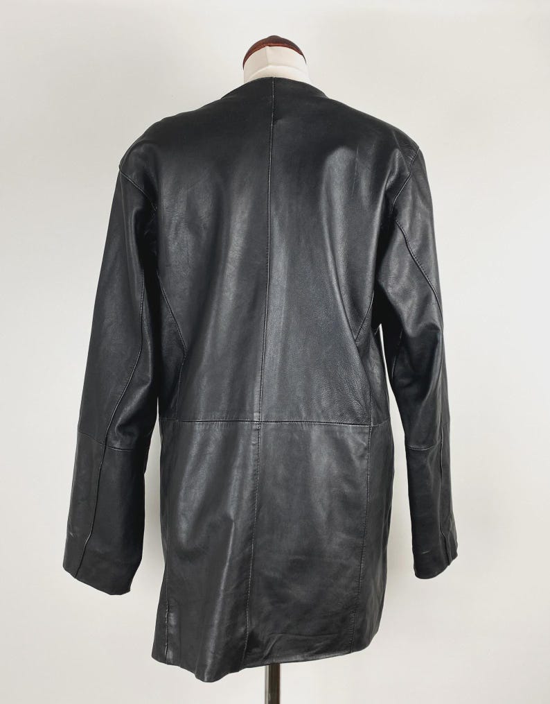 May include: A black leather coat displayed on a mannequin. The coat has a classic design with a collarless neckline, long sleeves, and a mid-thigh length. The leather appears smooth and has visible stitching details.