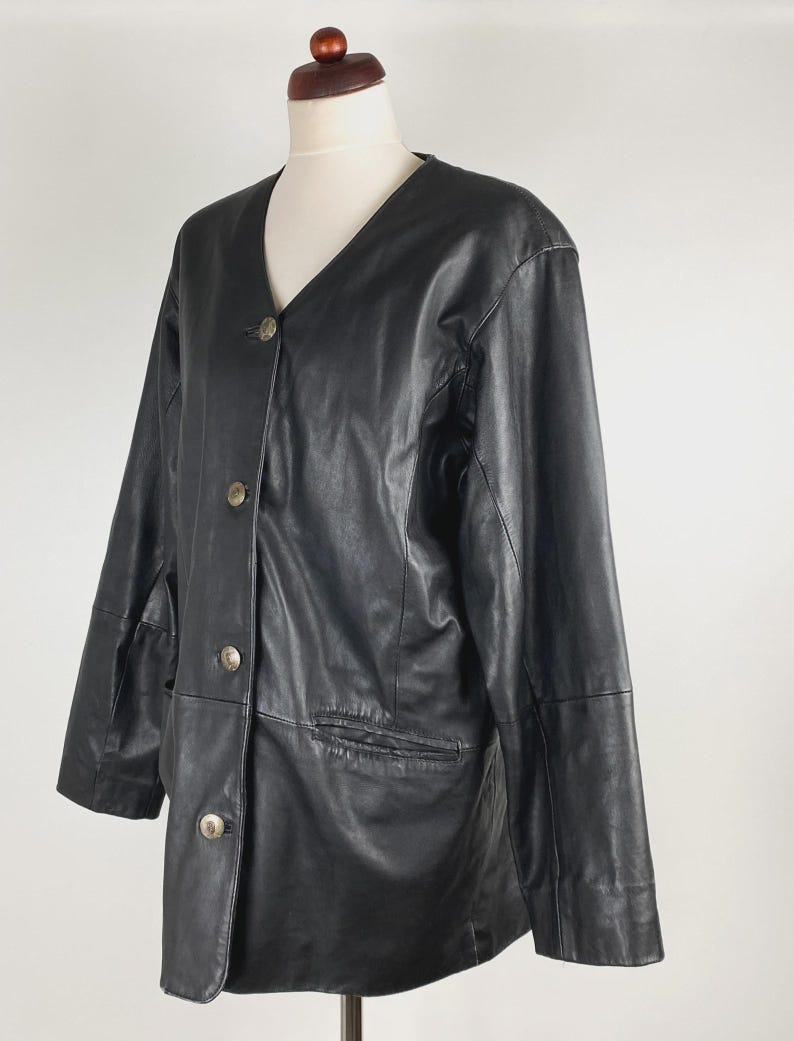 May include: Black leather jacket with a V-neckline and three silver-toned buttons. The jacket has two front pockets and is displayed on a mannequin. The jacket is a classic style.