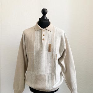 Puede incluir: Polo beige de manga larga con un patrón texturizado. La camisa presenta un cuello, tres botones marrones y un pequeño parche cuadrado en el pecho. La prenda se exhibe en un maniquí negro.