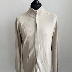 Könnte beinhalten: Hellbeige Reißverschluss-Pullover mit hohem Kragen. Der Pullover hat lange Ärmel und ein kleines Logo auf der Brust. Der Reißverschluss verläuft mittig am Pullover. Der Pullover wird auf einer Schaufensterpuppe präsentiert.