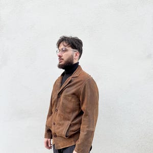 K&ouml;nnte beinhalten: Braune Wildlederjacke mit klassischem Kragen und Knopfverschluss. Die Jacke hat zwei Vordertaschen und wird &uuml;ber einem schwarzen Rollkragenpullover getragen. Die Person tr&auml;gt eine schwarze Hose und eine Brille.