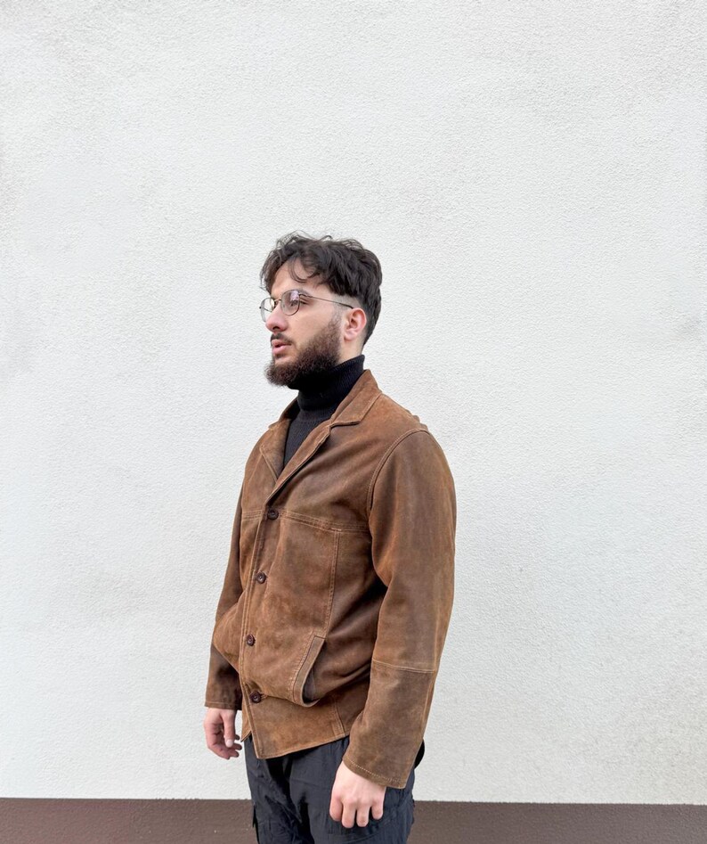 K&ouml;nnte beinhalten: Braune Wildlederjacke mit klassischem Kragen und Knopfverschluss. Die Jacke hat zwei Vordertaschen und wird &uuml;ber einem schwarzen Rollkragenpullover getragen. Die Person tr&auml;gt eine schwarze Hose und eine Brille.