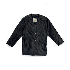 May include: Black leather jacket with a V-neckline and three gold-colored buttons. The jacket has long sleeves and two front pockets. A small tag is visible near the neckline. The jacket is laid flat on a white surface.