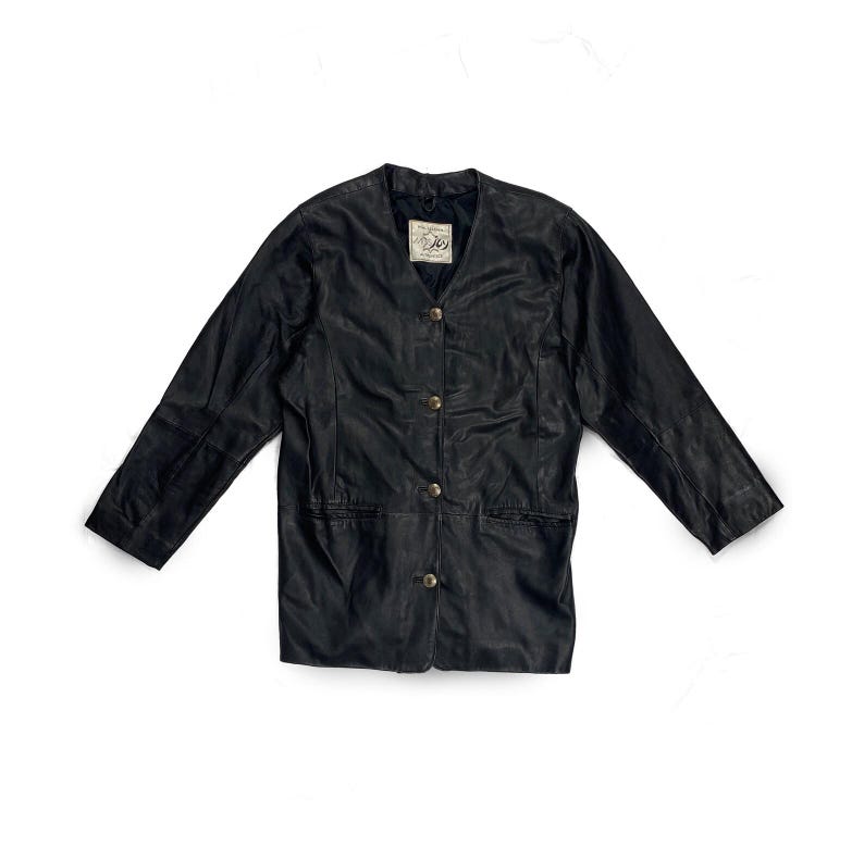 May include: Black leather jacket with a V-neckline and three gold-colored buttons. The jacket has long sleeves and two front pockets. A small tag is visible near the neckline. The jacket is laid flat on a white surface.