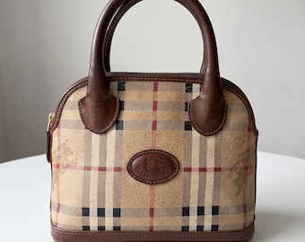 May include: A dome-shaped handbag featuring the iconic Burberry tan, black, and red check pattern. The bag has dark brown leather handles and trim, a gold-tone zipper, and a central oval leather patch embossed with the Burberry knight logo.