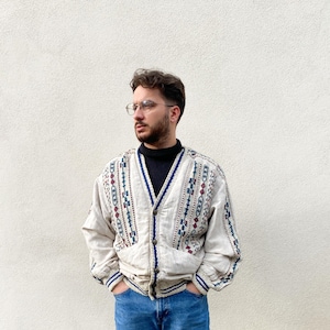 Op de afbeelding: Beige vest met een V-hals, met een geometrisch patroon in blauw, rood en groen. Het vest heeft een knoopsluiting, twee zakken en blauwe biezen. De persoon draagt een zwarte coltrui en een blauwe jeans.