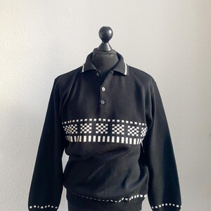 Könnte beinhalten: Schwarzer Langarmpullover mit Polokragen und drei Knöpfen. Der Pullover hat ein weißes und schwarzes Karomuster auf der Brust und weiße Verzierungen an Kragen, Manschetten und Saum. Das Kleidungsstück wird auf einer schwarzen Schaufensterpuppe präsentiert.