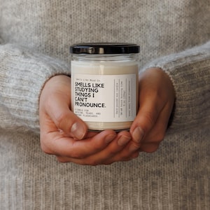 May include: A clear glass jar candle with a black lid, held in both hands. The candle has a white label with the text "Smells Like Studying Things I Can't Pronounce." The candle is a soy candle and has a 50-60 hour burn time.