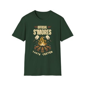 May include: Dark green t-shirt with a graphic featuring a campfire, marshmallows, and the text "OFFICIAL S'MORES TASTE TESTER". The design includes a campfire with flames and logs, and two marshmallows on sticks.