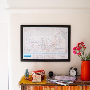 May include: A black-framed map of a subway system is mounted on a wall. A wooden shelf below displays a red vase with pink flowers, a globe, a clock, and other decorative items. The map is a subway map.