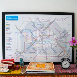 Puede incluir: Un mapa enmarcado del sistema de transporte público S-Bahn y U-Bahn de Berlín, con un marco negro. Un juguete de autobús de dos pisos rojo, un pequeño juguete de robot, un libro y un despertador negro están sobre una mesa frente al mapa. Un jarrón de flores está a la derecha.