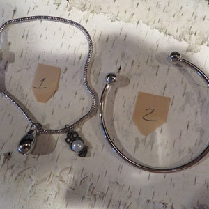 May include: Two silver bracelets, one with a charm of a bird and a teardrop, and the other with a round charm. The bracelets are on a white background with numbers 1 and 2 written on pieces of paper.