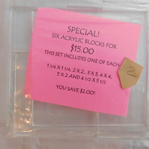 Puede incluir: Una nota rosa con el texto "SPECIAL! SIX ACRYLIC BLOCKS FOR $15.00 THIS SET INCLUDES ONE OF EACH: 1 1/4 X 1 1/4, 2 X 2, 3 X 3, 4 X 4, 5 X 2 AND 4 1/2 X 5 1/2 YOU SAVE $2.00!" La nota est&aacute; encima de un contenedor de pl&aacute;stico transparente con seis compartimentos.