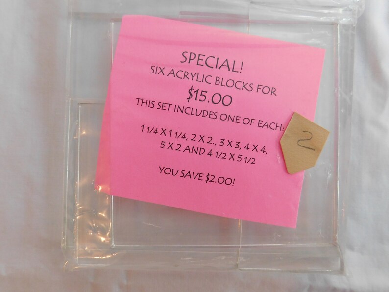 Puede incluir: Una nota rosa con el texto "SPECIAL! SIX ACRYLIC BLOCKS FOR $15.00 THIS SET INCLUDES ONE OF EACH: 1 1/4 X 1 1/4, 2 X 2, 3 X 3, 4 X 4, 5 X 2 AND 4 1/2 X 5 1/2 YOU SAVE $2.00!" La nota est&aacute; encima de un contenedor de pl&aacute;stico transparente con seis compartimentos.