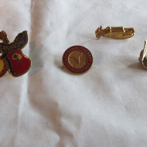 May include: Four gold-colored pins with different designs. The first pin features a banjo and guitar with a red and black banner that says "Branson". The second pin is a round red and gold pin with the words "Occupational Therapy Assistant". The third pin is a gold-colored airplane. The fourth pin is a small American flag.
