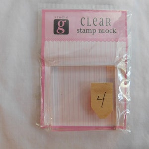 Puede incluir: Bloque de sello transparente de pl&aacute;stico en un embalaje rosa y blanco con el texto "studio g CLEAR stamp block" y el n&uacute;mero "4" escrito en una peque&ntilde;a etiqueta marr&oacute;n.