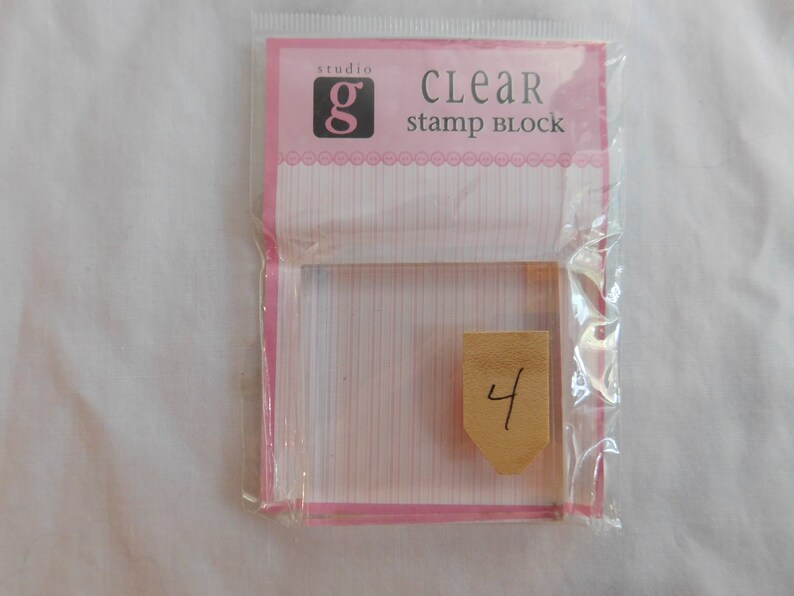 Puede incluir: Bloque de sello transparente de pl&aacute;stico en un embalaje rosa y blanco con el texto "studio g CLEAR stamp block" y el n&uacute;mero "4" escrito en una peque&ntilde;a etiqueta marr&oacute;n.