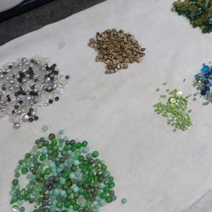 May include: A collection of beads in diverse colours and sizes, including clear, silver, brown, green, and blue. The beads are spread on a white cloth, highlighting their different shapes and textures. Suitable for jewellery making or craft projects.