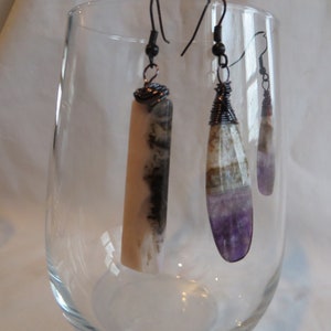 May include: Three pairs of gemstone earrings with dark metal hooks. The earrings feature rectangular and teardrop-shaped stones in shades of white, black, brown, and purple. The stones are wrapped with wire.