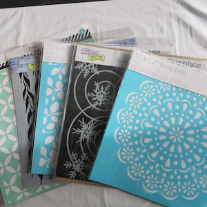 May include: Five 12x12 inch stencils for crafting. The stencils are made of clear plastic and feature various designs, including snowflakes, leaves, and a circular pattern. The stencils are packaged in clear plastic sleeves.