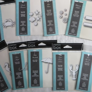 May include: A collection of eight metal craft dies packaged in clear plastic bags. The dies are for use on cardstock, felt, fabric, and shrink plastic. The dies feature various designs, including a crab, a seahorse, a lobster, an umbrella, a hot air balloon, a swirl, a label, and gears.